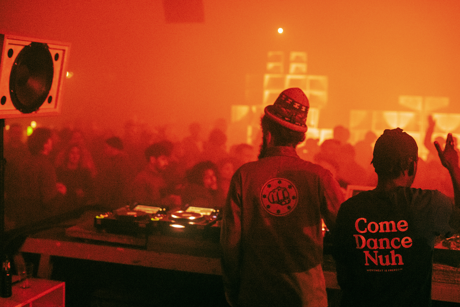 This photo has an orange colour as its base. The photo is taken from the DJ booth. You see Errol & Sammseed from the back. In front of them you see people dancing. On right side, all the way in the back, you see KrackFree's soundsystem.